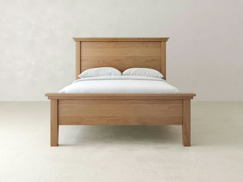 The Ivy Bed from James+James showcases a wooden frame complete with a matching headboard and footboard, capturing a minimalist yet timeless aesthetic. Adorned with crisp white sheets and two pillows, the bed is placed against a simple, light-colored wall on a pristine white floor.