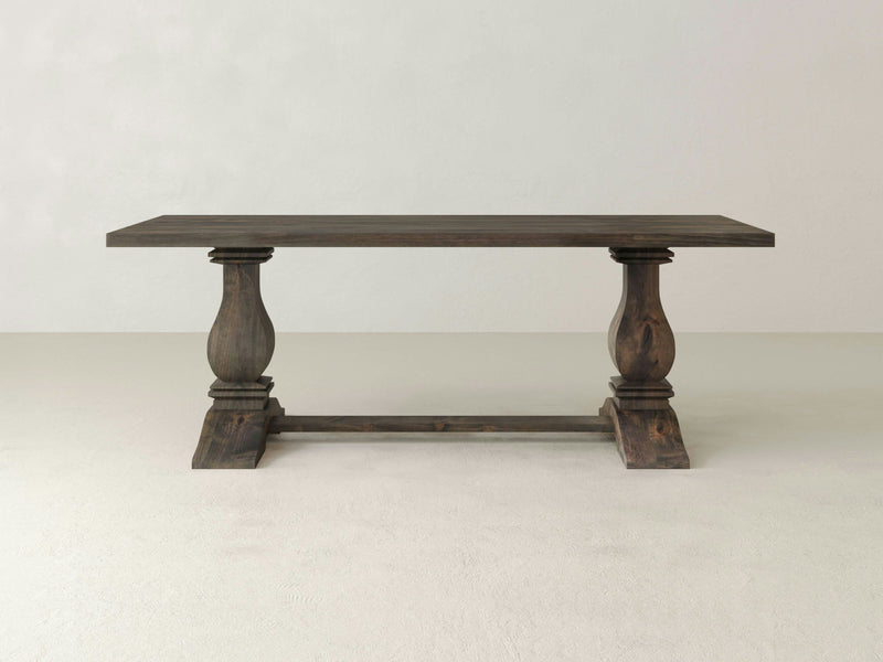 The Heirloom Dining Table - Deep Grey by James+James features a dark wooden rectangular top supported by two ornate, carved legs connected by a horizontal stretcher, all set against a plain, light-colored background. The table boasts a classic and sturdy design.