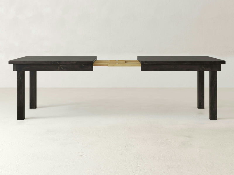 The Farmhouse Expandable Dining Table - Charred Ember by James+James features a dark wooden, rectangular top and four sturdy legs. A visible seam down the middle highlights its extendable design, ideal for accommodating more people or items. The background is plain white.