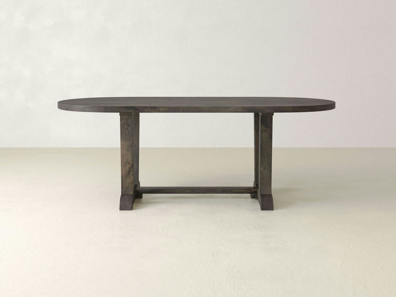 The Thaden Oval Dining Table - Deep Grey by James+James features a minimalist, dark wooden design with a simple, sturdy construction. The table is supported by two legs on either end, connected by a horizontal stretcher. It is displayed on a light, neutral-colored floor against a plain, off-white background.