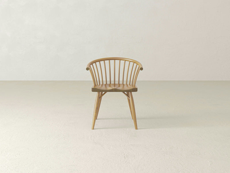 A Maria Dining Chair - Harvest Wheat by James+James, featuring a wooden construction with a curved backrest and vertical slats, is situated on a light-colored floor against a plain, light-colored wall. The chair boasts a minimalist design with tapered legs and a smooth finish.