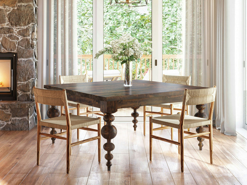 A cozy dining area with the Olivia Square Dining Table - Tobacco by James+James and four wicker chairs. A glass vase with white flowers sits on the table. The room features large windows with sheer curtains, a stone fireplace on the left, and a view of a wooden deck and greenery outside.