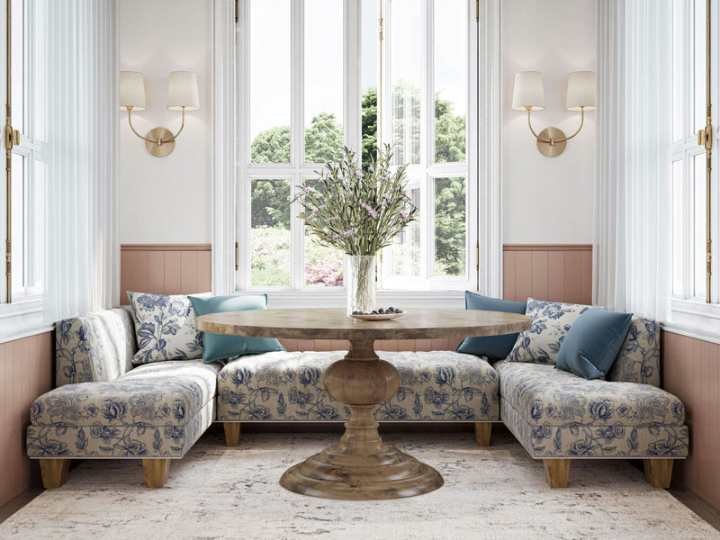 A cozy corner nook features the Josephine Round Dining Table in Harvest Wheat by James+James, surrounded by L-shaped upholstered benches adorned with blue floral patterns and matching blue cushions. Tall windows with white frames allow natural light to flood the space, while wall sconces add an elegant touch.