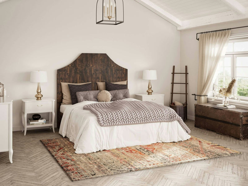 A cozy bedroom features the Megan Bed in Tobacco by James+James, adorned with white bedding and patterned throw pillows. The room includes bedside tables with lamps, a vintage trunk, an antique ladder, a large window with sheer curtains, and a patterned rug on parquet flooring.