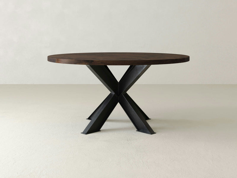 A Shiloh Round Dining Table - Tobacco by James+James, featuring a dark brown top and black X-shaped metal legs, is placed in a minimalistic room with a light-colored floor and wall.