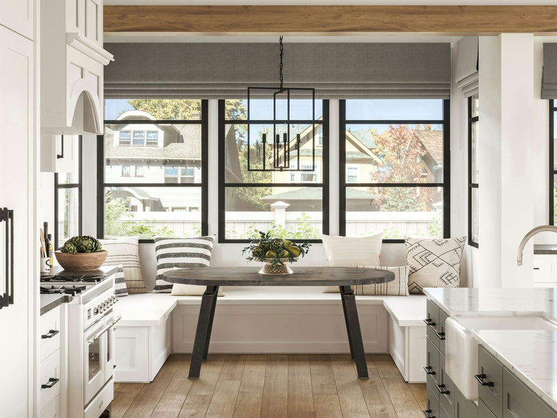 A modern kitchen with white cabinetry, stainless steel appliances, and a large island with a sink. The space features a Fulton Oval Dining Table in Deep Grey by James+James, adorned with a centerpiece and flanked by a bench and chairs. The dining area is set by a window nook with tall windows letting in natural light.