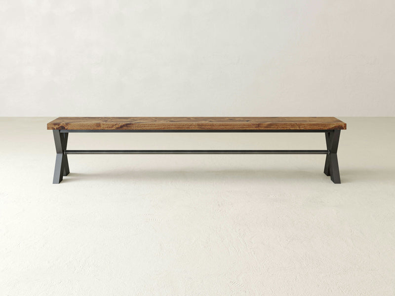 The X-Base Bench - Tuscany by James+James stands against a plain, light-colored background. This long, minimalist wooden bench features a dark iron base and boasts a natural wood grain finish with a simple, sturdy design, offering a modern yet rustic aesthetic.