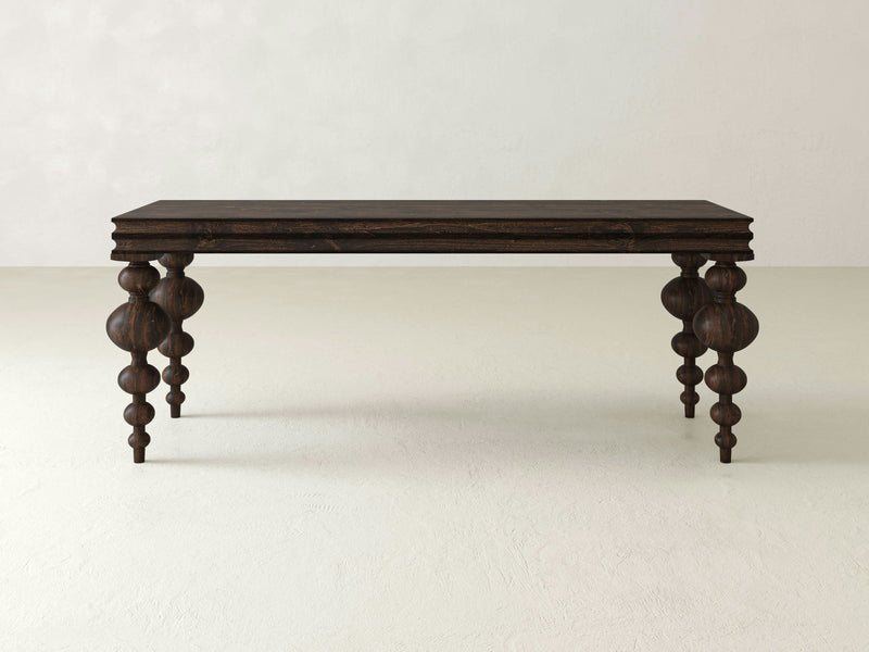 The Olivia Sofa Table - Tobacco by James+James, featuring a dark wooden finish and four intricately carved, spindle-style legs, stands against a plain, light-colored background. The table boasts a rectangular top with a simple, flat surface.