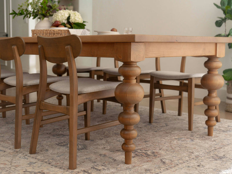 The Olivia Expandable Dining Table - Harvest Wheat by James+James, featuring intricately carved legs, is complemented by four matching chairs with cushioned seats. The table is adorned with a basket of flowers and set on a light-patterned rug in a bright room, with a small potted plant in the background.