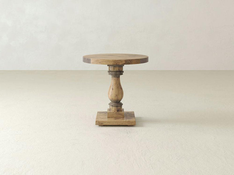 The Vivien Side Table - Harvest Wheat by James+James stands on a light-colored floor against a plain, light-colored wall. This rustic wooden pedestal table boasts a round top, a turned central support, and a square base. The natural and lightly weathered wood finish enhances its vintage charm.