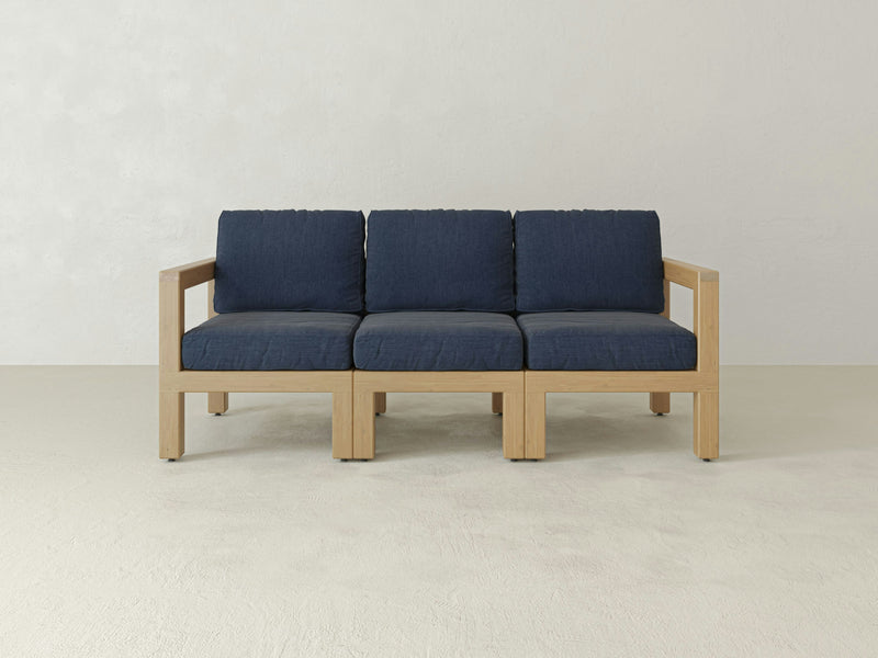 The James+James Aria Outdoor Sofa, characterized by its wooden frame and three navy blue padded cushions, is elegantly placed in a minimalistic setting against a plain, light-colored wall. This piece highlights clean lines and a simple aesthetic while offering customizable features perfect for outdoor living.