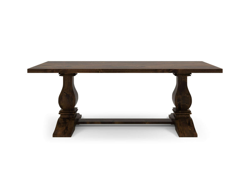 The Heirloom Dining Table - Tobacco by James+James is a dark wooden dining table featuring a rectangular top and two intricately carved pedestal legs connected by a horizontal support beam. It boasts a classic, rustic design with a smooth, polished finish.