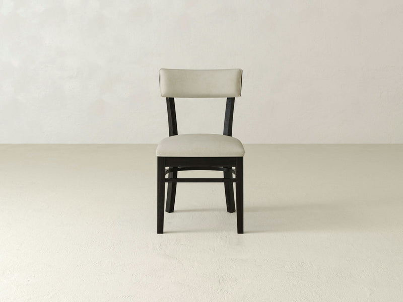 The Everly Side Dining Chair - Charred Ember by James+James is a simple, modern chair featuring a light beige cushioned seat and backrest. Its frame and legs are crafted from dark wood. The chair is positioned against a plain, off-white wall and sits on a light grey floor.