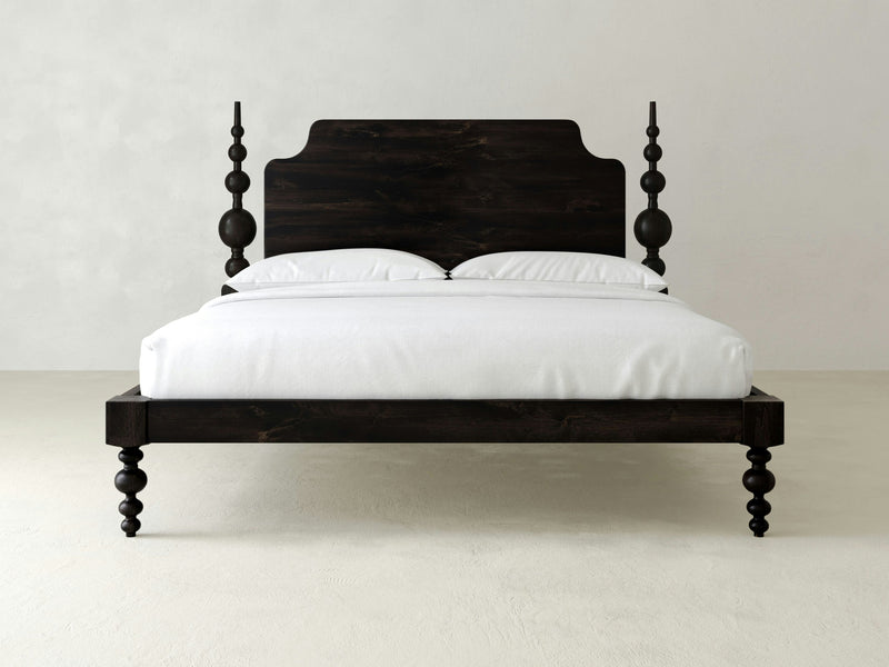 The Opal Bed - Charred Ember by James+James features a dark, polished wooden finish. The bed showcases a high, ornate headboard and matching foot posts. It is neatly made with white bedding, including two pillows and a duvet. The background has a plain, light-colored wall and floor.