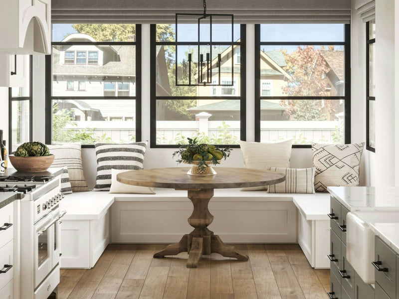 A cozy kitchen nook featuring the Sybil Round Dining Table in Harvest Wheat by James+James is surrounded by built-in white bench seating adorned with patterned cushions. Large windows provide ample natural light, showcasing a view of neighboring houses. A modern black chandelier hangs above the table.