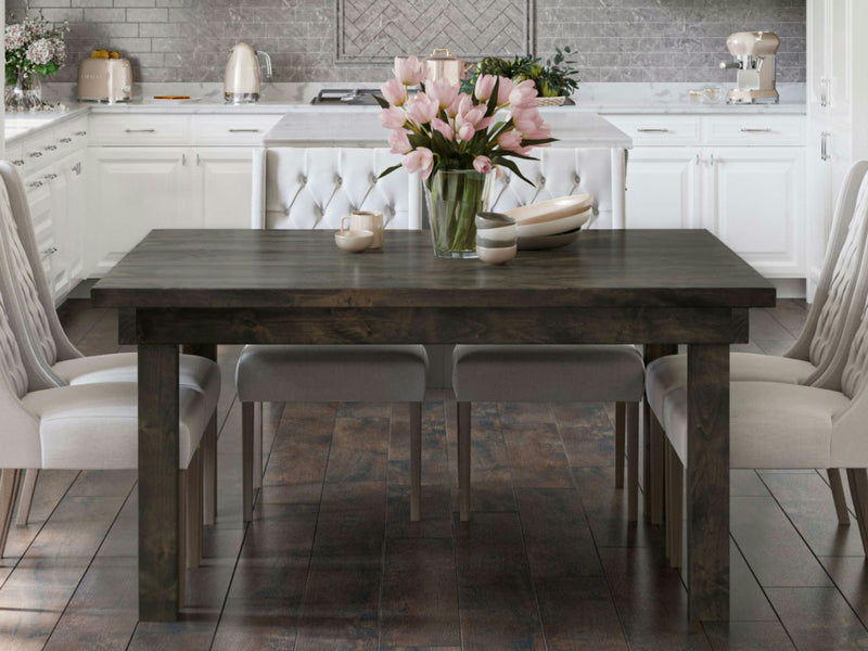 A modern kitchen with white cabinets and grey accents features a Farmhouse Square Dining Table in deep grey from James+James at the center. The table is set with bowls, plates, a teacup, and a vase with pink tulips. Four light grey cushioned chairs surround the table, and various kitchen appliances are visible in the background.