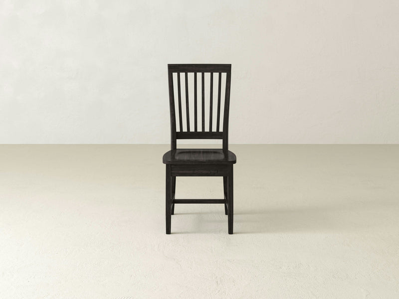 The James+James Jane Dining Chair, characterized by its single dark wooden frame and vertical slats on the backrest, is displayed on a light-colored floor against a plain off-white background. Ideal for any dining setting, its simple and classic design includes a flat seat and four sturdy legs. Customizable stain options are available to suit your preferences.