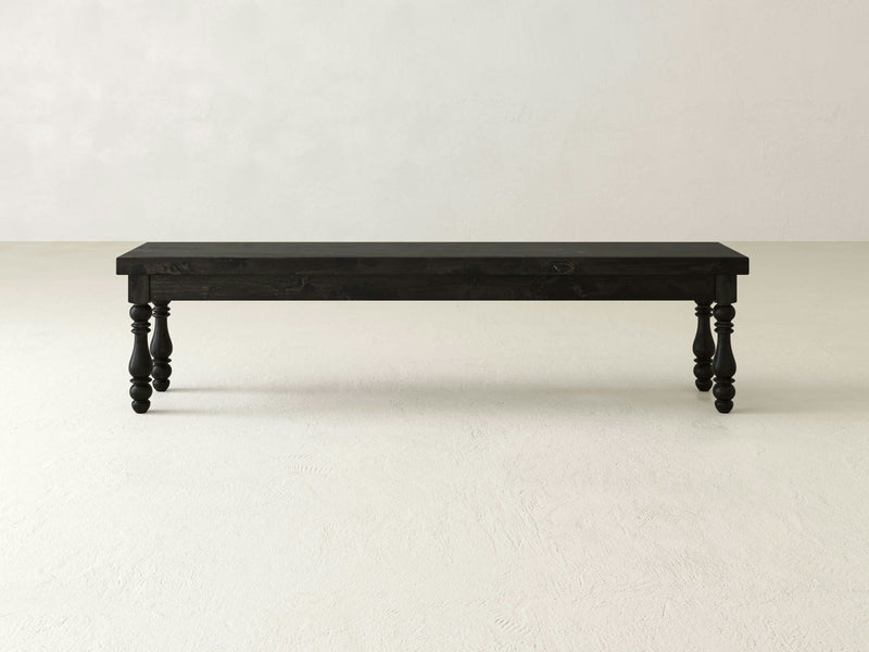 The Annli Bench by James+James exudes dining elegance with its minimalist wooden design, featuring a dark finish, rectangular seat, and four turned legs. It rests on a light-colored floor against a subtly textured wall in the neutral space, embodying both sophistication and robust design.