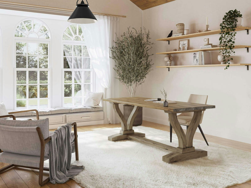 A bright, modern room with large arched windows letting in natural light. The space features a Vera Desk by James+James in a Harvest Wheat finish paired with a chair, a cozy window seat with cushions, a plush white rug, an armchair, and minimalist wall shelves holding books, decor, and a trailing plant.