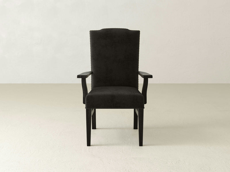 A single black Hadley Arm Dining Chair from James+James, upholstered and centrally positioned against a neutral, light-colored wall background. The chair features a high backrest and studded detailing along the edges, with wooden legs extending to the floor. This piece exudes sophistication, perfect for an elegant dining experience.