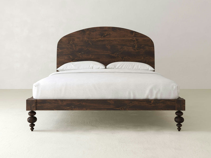 An Eden Bed - Tobacco by James+James, featuring an arched headboard and four turned legs, stands against a plain white background. The bed is dressed with a white fitted sheet and two white pillows, giving a minimalist and clean appearance.