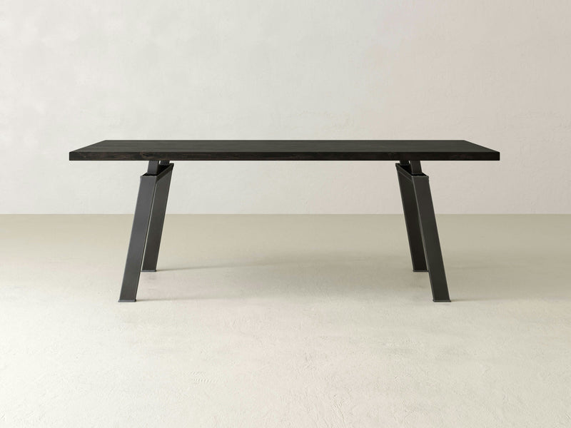 The Fulton Dining Table - Charred Ember by James+James boasts a minimalist, modern design featuring a rectangular dark wooden top and angled black metal legs, set against a plain, light-colored background. Its clean lines and sleek appearance ensure it fits seamlessly into any contemporary space.