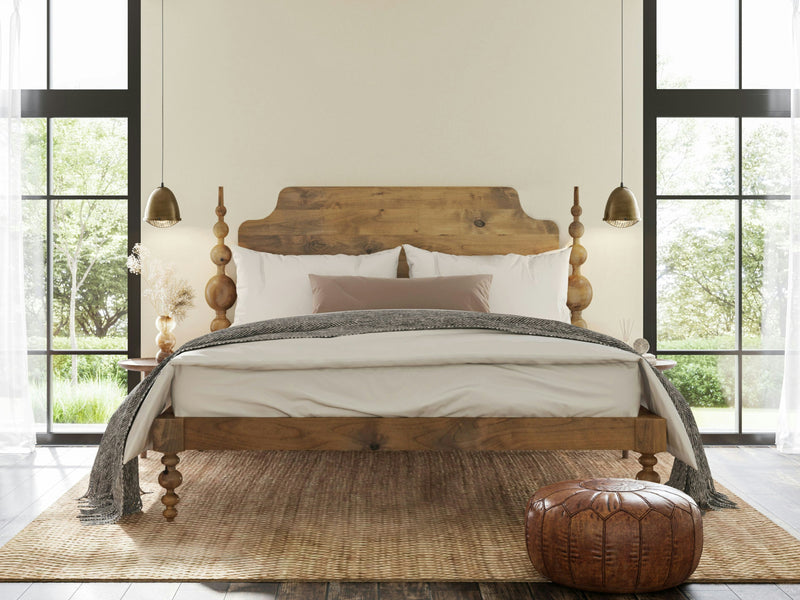 A cozy bedroom features the Opal Bed in Harvest Wheat by James+James, with its elegant turned posts and rustic headboard, adorned with white and gray bedding. Windows with black frames let in natural light, flanking the bed. Pendant lights hang above the bedside tables, and a round pouf sits on the woven rug.