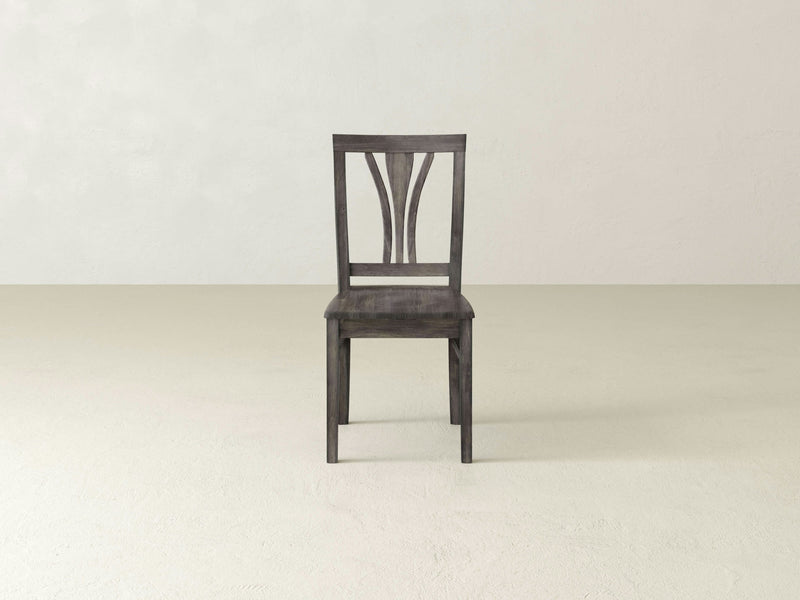 The Brooklyn Dining Chair - Deep Grey by James+James is the focal point of the image, standing on a light-colored floor against a plain off-white wall. The chair features a simple backrest design and clean lines, with a sturdy appearance. It's centered in the image, showcasing its minimalist elegance.