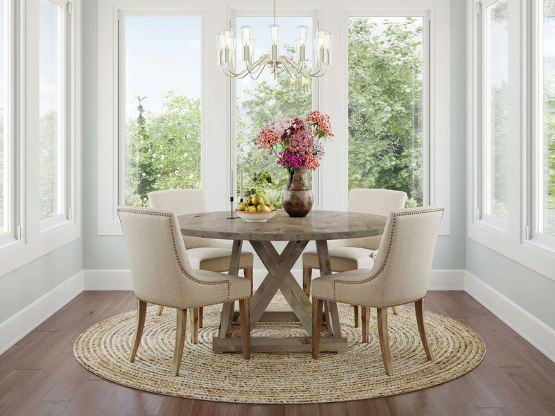 A bright dining room with large windows features the Julia Round Dining Table in Barn Wood by James+James. Four beige upholstered chairs are arranged around the table, which is topped with a vase of pink and purple flowers and a bowl of fruit. The table sits on a round woven rug, and a chandelier hangs above.