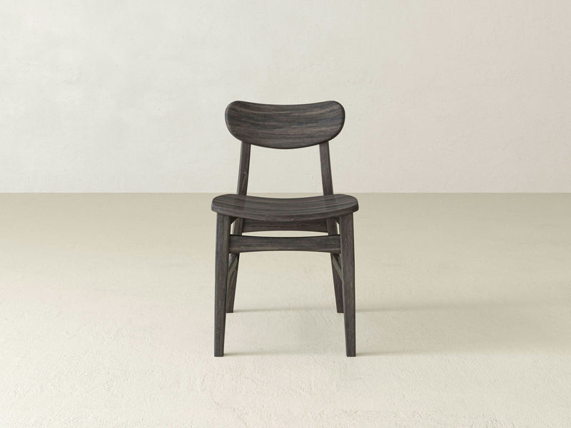 The Lynn Dining Chair - Barn Wood by James+James is a wooden chair with a curved backrest and seat, viewed from a rear angle. The chair boasts a dark finish and simple, sturdy construction featuring four legs and a support structure connecting them at the base. Its design is both minimalistic and functional.