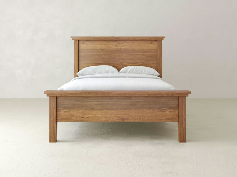 An Ivy Bed - Harvest Wheat by James+James, featuring a headboard and footboard, is beautifully adorned with two pillows and a neatly made white bedspread. It stands centered against a plain, light-colored wall in a minimalistic room.