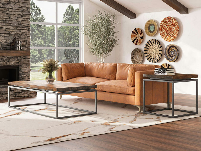 A modern living room with a brown leather sofa, a large rectangular Floating Top Coffee Table - Tuscany by James+James, and a matching side table. The wall features decorative woven baskets. A large window allows natural light in, and there's a stone fireplace on the left with green trees outside.