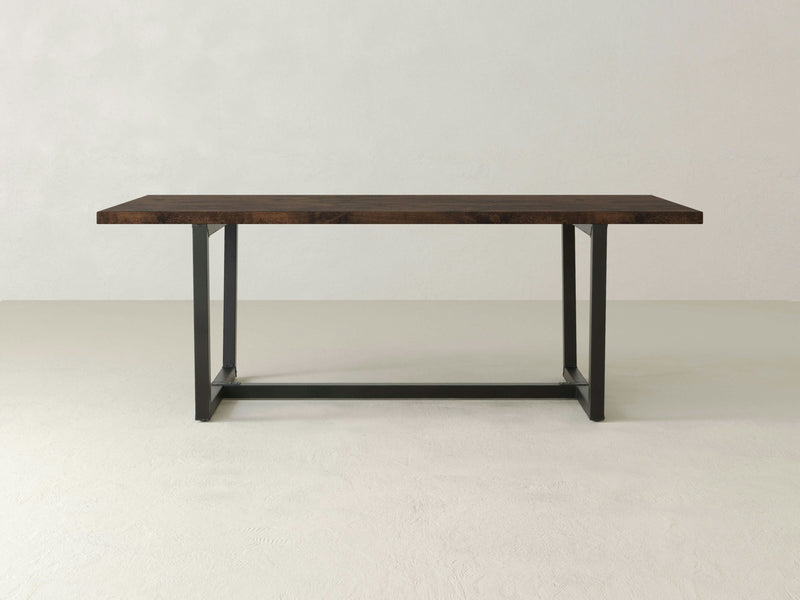 The Trapezoid Dining Table - Tobacco by James+James is a minimalist wooden table featuring a dark brown rectangular top and a black metal frame base, standing on a light-colored floor against a plain off-white background.