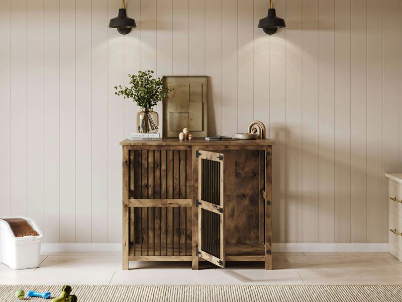 A cozy interior with a wooden cabinet against a light-colored, paneled wall. The cabinet has open slatted doors and decorative items, including a vase with greenery and a framed picture on top. Two wall-mounted lights illuminate the scene, while the Single Dog Kennel - Harvest Wheat by James+James sits nearby.