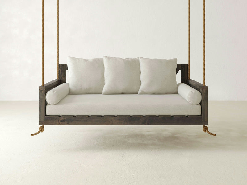 A minimalist scene showcasing the Avery Porch Swing - Deep Grey by James+James, suspended by sturdy ropes. The swing features a dark wooden frame with light gray cushions, including three square back cushions and two cylindrical side bolsters. A plain and neutral background enhances the calm ambiance.