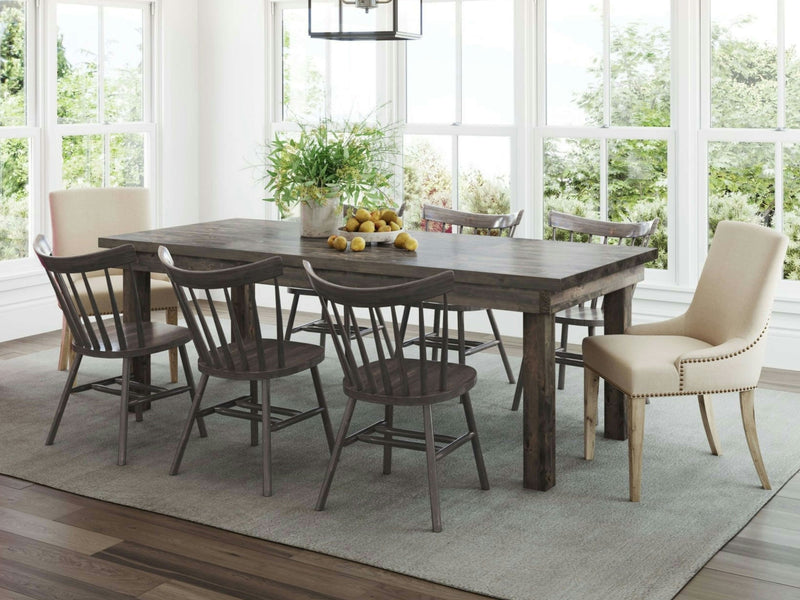 A dining room featuring the Farmhouse Dining Table - Deep Grey by James+James, surrounded by an eclectic mix of chairs, including beige upholstered end chairs and dark wooden side chairs. A centerpiece with lush green plants and a bowl of lemons decorates the table, while large windows provide a scenic view of greenery outside.