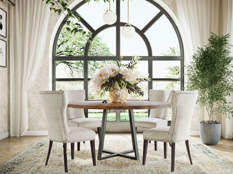 A dining area featuring the Watson Round Dining Table in Tuscany by James+James is surrounded by four upholstered chairs. A floral centerpiece adorns the table. The backdrop showcases a large arched window with lush greenery outside, framed by light-colored curtains that complement a nearby potted plant.