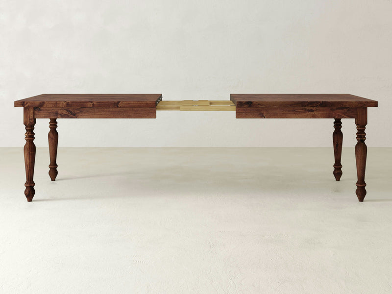 The James+James French Country Expandable Dining Table features a rich, dark wood top and ornate, turned legs. With its extension partially pulled out, the mechanism and additional leaves are revealed. The rustic beauty of this piece is highlighted against a plain, light-colored wall and floor.