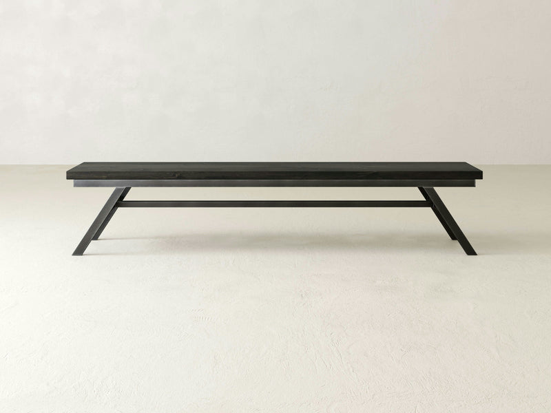 The Fulton Bench - Charred Ember by James+James is a modern, minimalist piece featuring a sleek, dark wooden seat paired with black metal legs. The legs are angled outward for enhanced stability and connected by a horizontal support bar that reinforces its sturdy appearance. This bench boasts a clean, contemporary design.