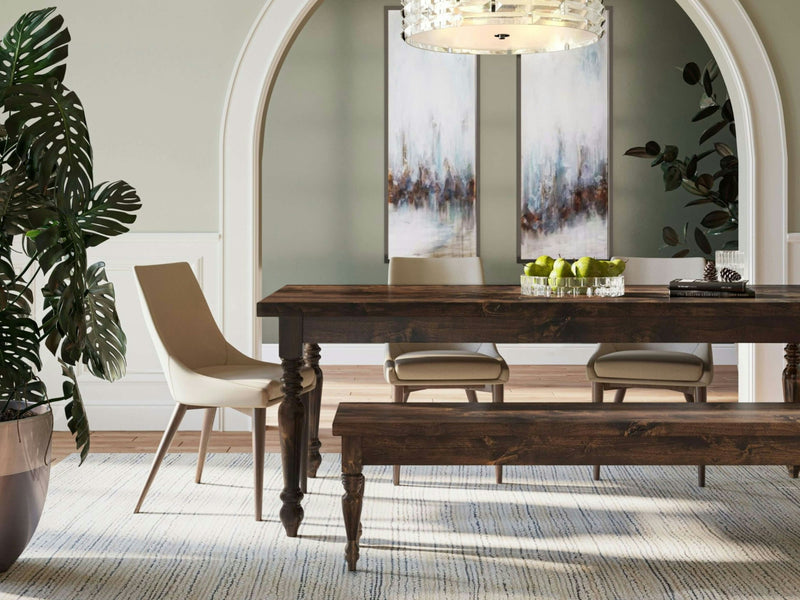 A dining room featuring the James+James French Country Expandable Dining Table in Tobacco, accompanied by a matching bench and flanked by light-colored chairs. A bowl of green apples decorates the table. The room boasts arched doorways, two abstract paintings on the wall, a large potted plant, and a modern chandelier suspended overhead.