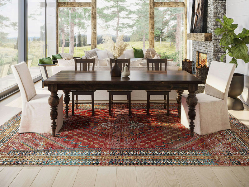 A spacious dining room features a large Bailey Expandable Dining Table in Tobacco by James+James, accompanied by six chairs—two with white slipcovers and four wooden. The room includes a large red patterned rug, floor-to-ceiling windows offering a forest view, and a fireplace to the right adorned with a nearby plant.