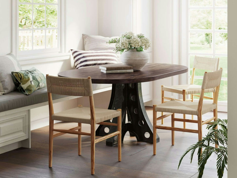 A cozy dining nook featuring the Amelia Oval Dining Table in Tobacco from James+James, surrounded by three wicker chairs and a cushioned bench by a sunlit window. A vase of white flowers sits on the table alongside a book, while a striped pillow and a leafy plant add decorative touches to the bright, airy space.