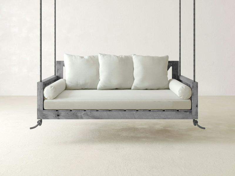 A minimalist light grey hardwood swing bed from James+James, featuring the Mattress Cushion and Pillows for Twin Swing adorned with durable beige Sunbrella cushions and two cylindrical bolster pillows, is suspended from the ceiling by thick ropes. The background of a plain white wall and floor enhances a serene and modern ambiance.