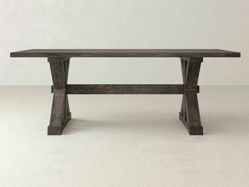 Introducing the Trestle Dining Table - Deep Grey by James+James: a simple, rustic wooden table featuring a rectangular top and sturdy X-shaped legs at each end. The craftsmanship of this dining table is beautifully accentuated by its dark, weathered finish, making it stand out against a plain, light-colored background.