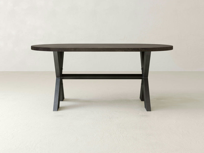 An empty, lightly textured room with a neutral-colored background features the X-Base Oval Dining Table - Deep Grey by James+James. This elegant, minimalistic wooden table has angled legs and a crossbar for added support, showcasing its sturdy construction and chic dark brown finish.