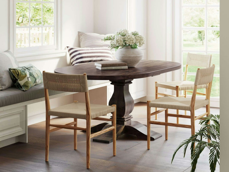 A cozy dining nook featuring the Heirloom Oval Dining Table in Tobacco by James+James, adorned with a vase of white flowers. The table is surrounded by two light-colored woven chairs and a built-in cushioned bench with striped and patterned pillows. Large windows in the background let in natural light.