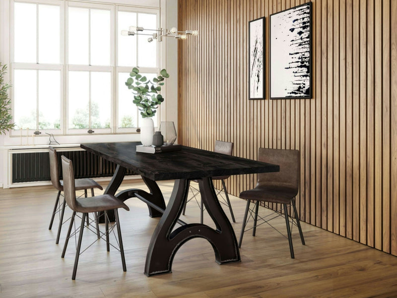 Modern dining room with the Wishbone Dining Table - Charred Ember by James+James, featuring industrial-style legs, surrounded by four brown chairs. A white vase with green foliage is centered on the table. The room has wooden paneling on one wall, large windows, and framed abstract art on the wall.