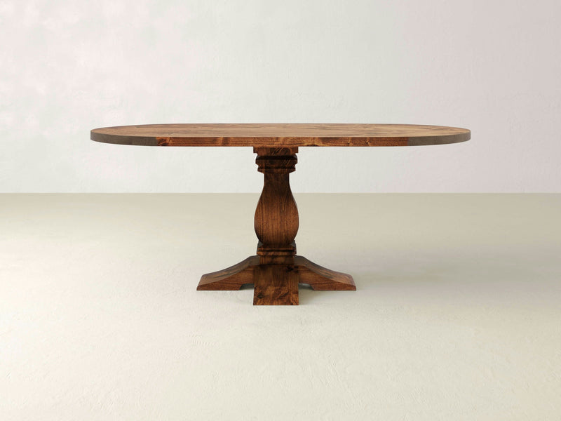 The Heirloom Oval Dining Table - Tuscany by James+James features a smooth surface and an intricately carved pedestal base. It stands on a light-colored floor against a plain background, showcasing its rich brown finish that highlights the natural wood grain.