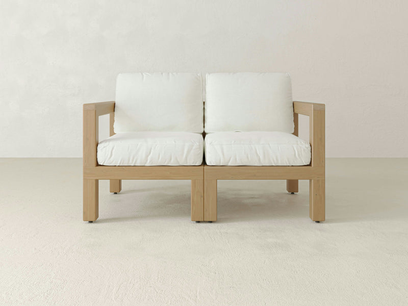 The Aria Outdoor Love Seat by James+James is a modern, minimalist piece featuring a wooden frame with green cushioned seats and backrests. This stylish outdoor loveseat boasts a clean, contemporary design and is set on a plain, light-colored floor against a simple, light-colored background.