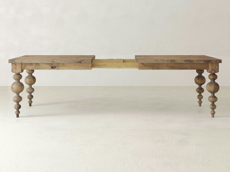 The Olivia Expandable Dining Table by James+James is a wooden dining table with a rustic finish and ornamental carved legs. Shown in its extended position, this solid hardwood table features a partially revealed leaf. The plain, light-colored wall and floor in the background accentuate its timeless design.